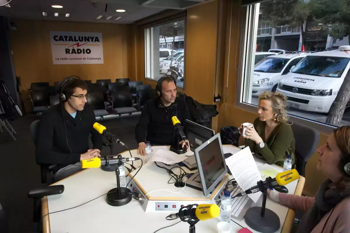 Cuatro personas están sentadas alrededor de una mesa en un estudio de radio con micrófonos y computadoras, mientras conversan y graban un programa en Catalunya Ràdio. Cuatro personas están sentadas alrededor de una mesa en un estudio de radio con micrófonos y computadoras, mientras conversan y graban un programa en Catalunya Ràdio.