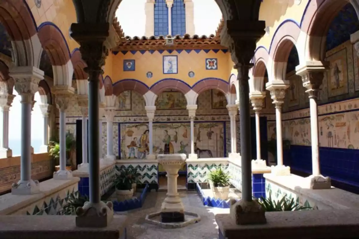 Patio interior con columnas y arcos decorados, paredes cubiertas de azulejos coloridos con escenas pintadas y una fuente central rodeada de plantas Patio interior con columnas y arcos decorados, paredes cubiertas de azulejos coloridos con escenas pintadas y una fuente central rodeada de plantas