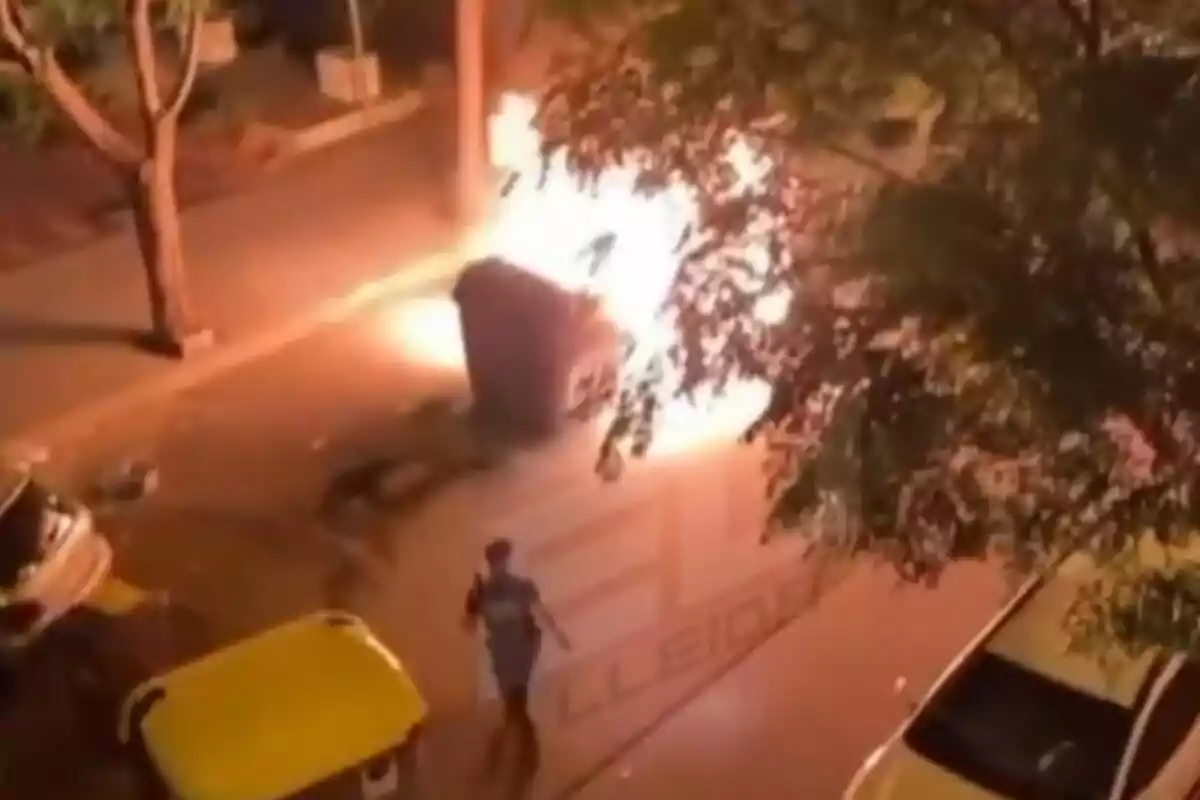 Contenedor de basura en llamas en una calle por la noche con una persona caminando cerca y varios árboles y autos alrededor