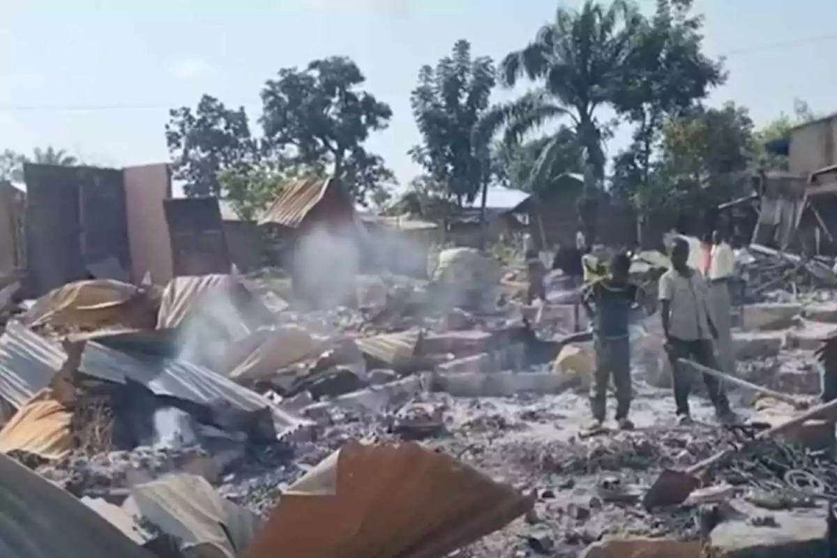 Personas de pie entre los restos humeantes de casas destruidas con árboles al fondo en un ambiente de devastación