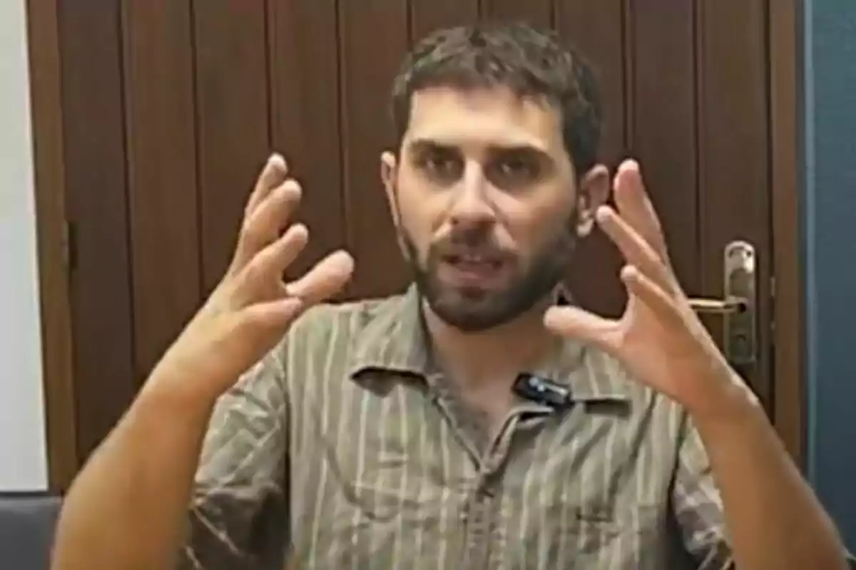 Hombre de barba y cabello corto gesticulando con ambas manos levantadas frente a una puerta de madera