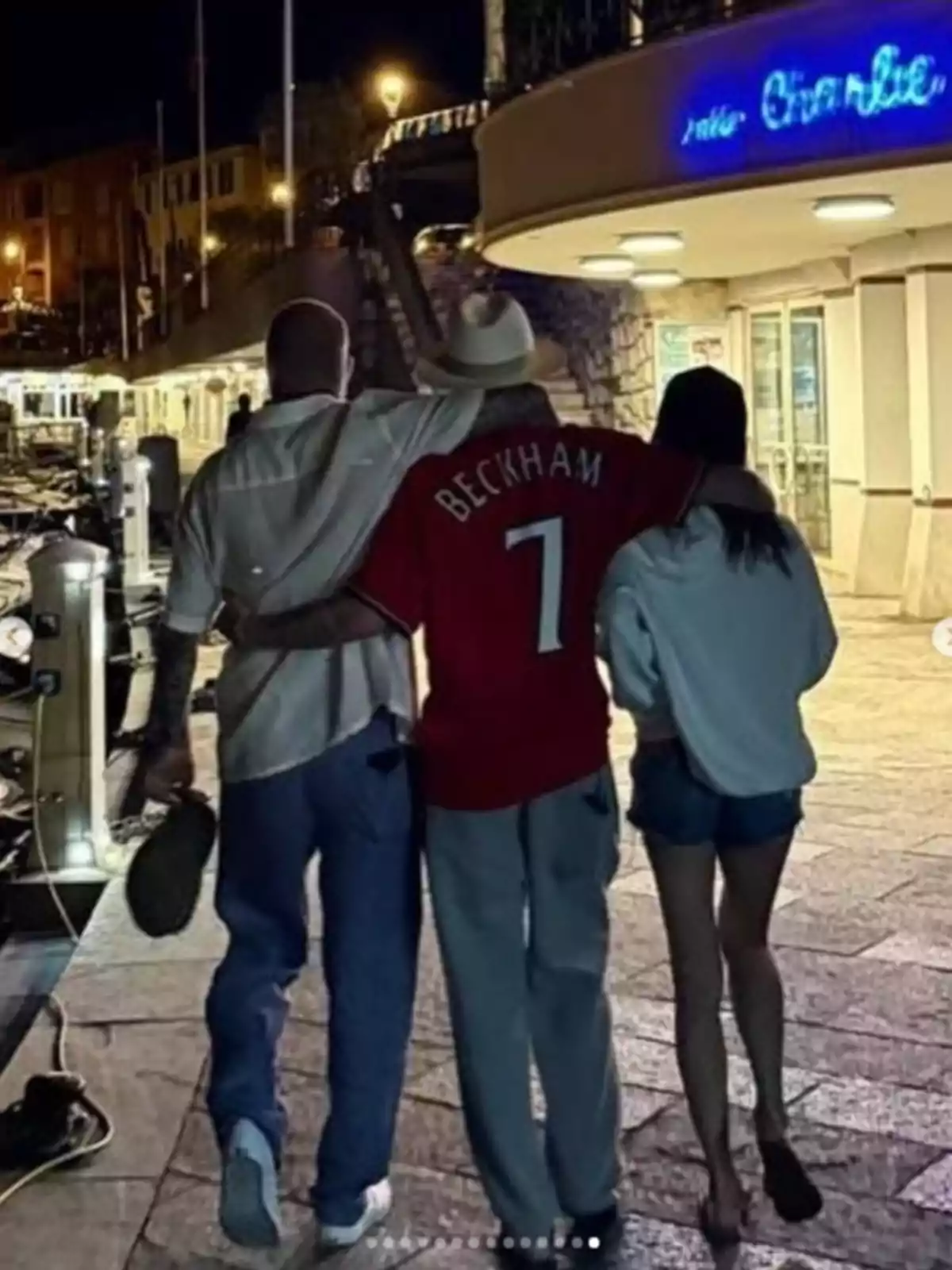 David, Victoria, and Cruz Beckham walking together from behind, the one in the middle wearing a red jersey with the name Beckham and the number 7, with their arms around the other two people on each side. David, Victoria, and Cruz Beckham walking together from behind, the one in the middle wearing a red jersey with the name Beckham and the number 7, with their arms around the other two people on each side.