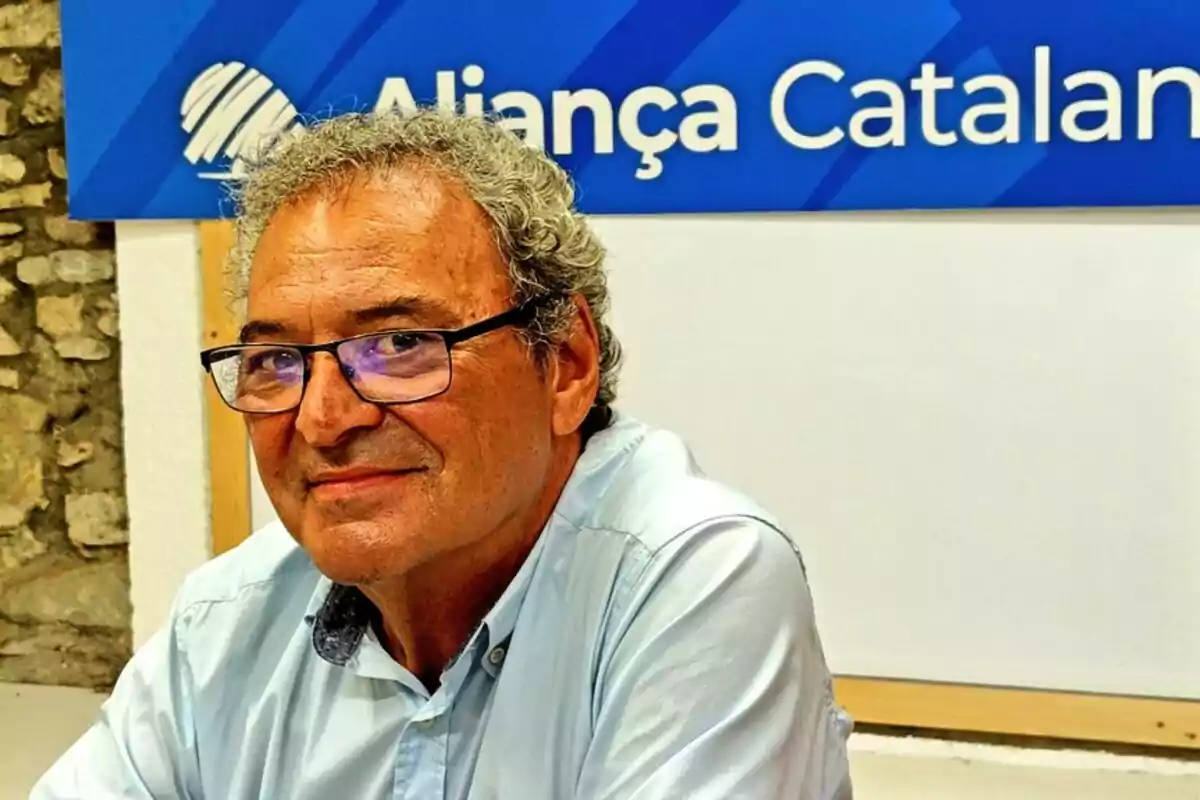 Home de cabells canosos i ulleres amb camisa clara assegut davant d’un cartell blau que diu Aliança Catalana