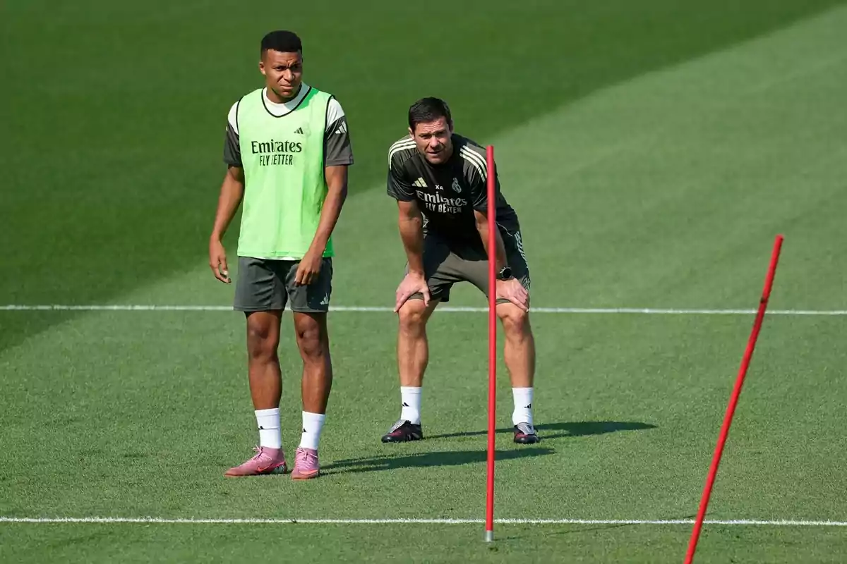 Dues persones amb roba esportiva del Reial Madrid són en un camp de futbol durant un entrenament al costat d’uns pals vermells. Dues persones amb roba esportiva del Reial Madrid són en un camp de futbol durant un entrenament al costat d’uns pals vermells.