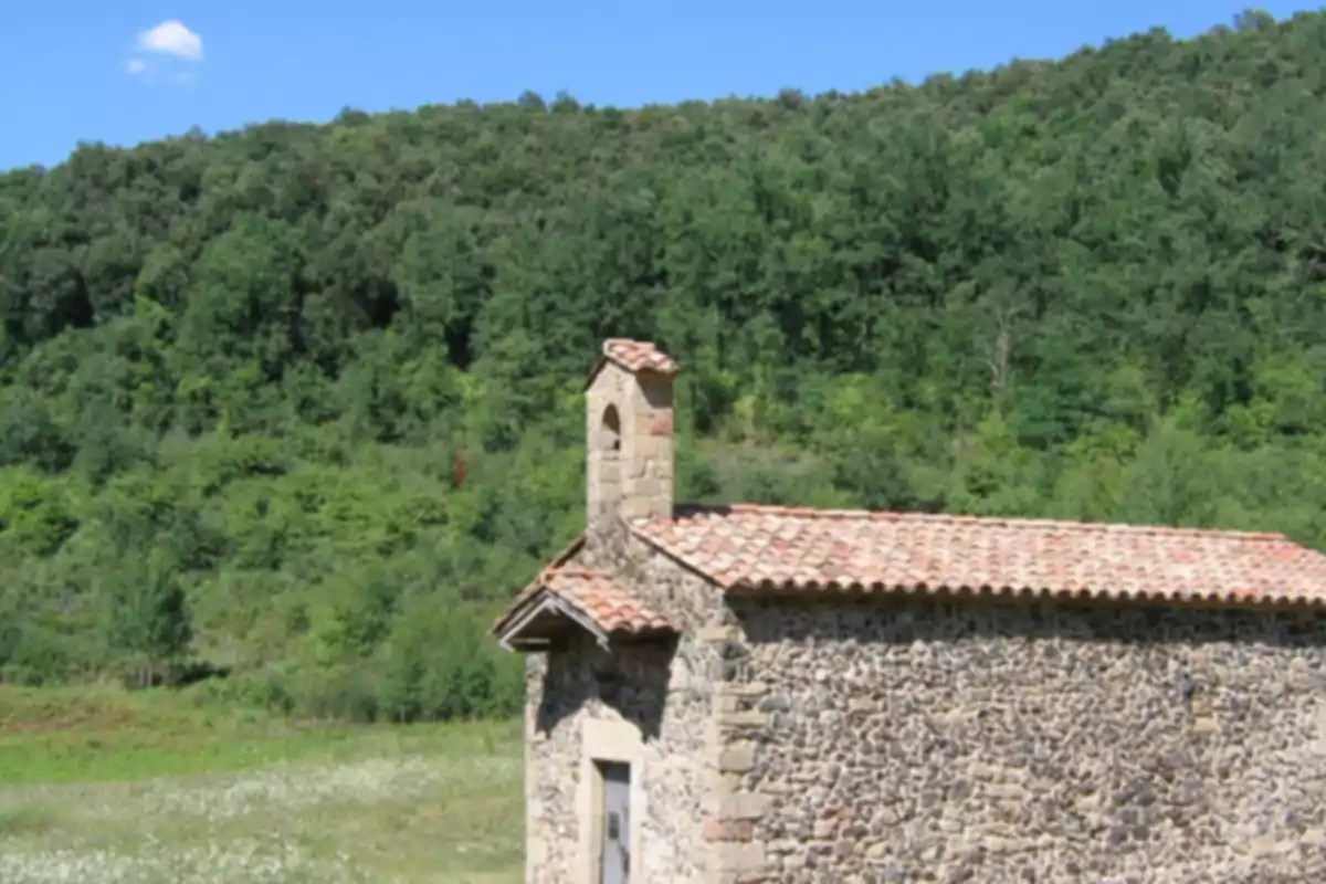Petita capella de pedra amb teulada de teules en un camp verd envoltat d’arbres i turons sota un cel blau Petita capella de pedra amb teulada de teules en un camp verd envoltat d’arbres i turons sota un cel blau