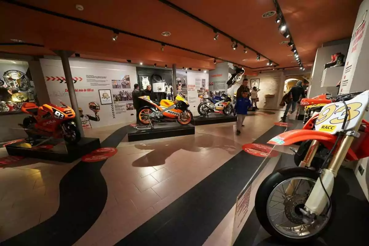 Exhibición de motocicletas deportivas en un museo con varias personas observando las motos y paneles informativos en las paredes Exhibición de motocicletas deportivas en un museo con varias personas observando las motos y paneles informativos en las paredes