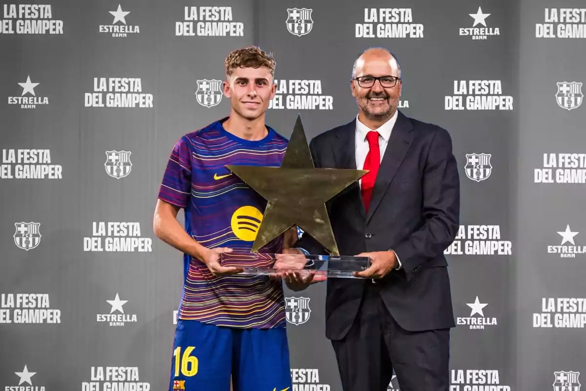Un futbolista del Barcelona i un home amb vestit sostenen junts un trofeu en forma d’estrella davant d’un fons amb el logotip de La Festa del Gamper. Un futbolista del Barcelona i un home amb vestit sostenen junts un trofeu en forma d’estrella davant d’un fons amb el logotip de La Festa del Gamper.
