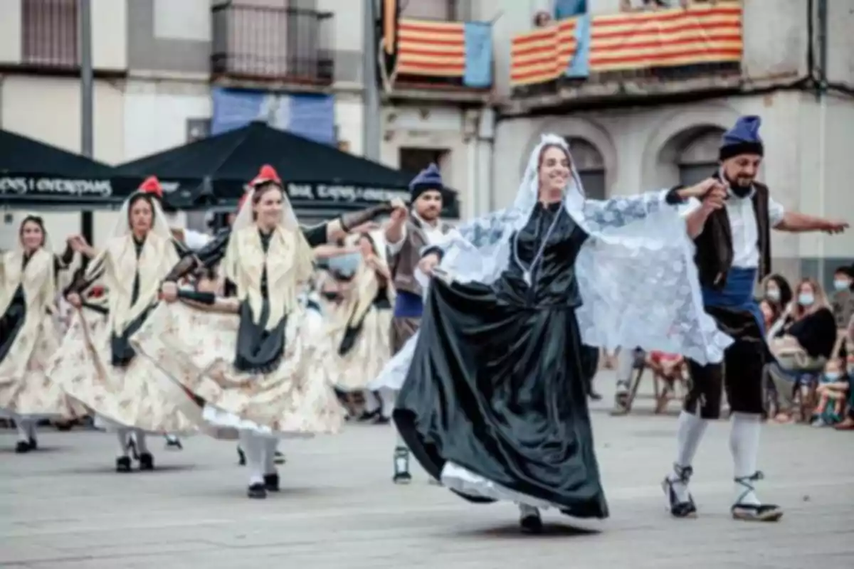 Personas bailando una danza tradicional con trajes típicos en una plaza decorada con banderas rayadas y público observando al fondo Personas bailando una danza tradicional con trajes típicos en una plaza decorada con banderas rayadas y público observando al fondo