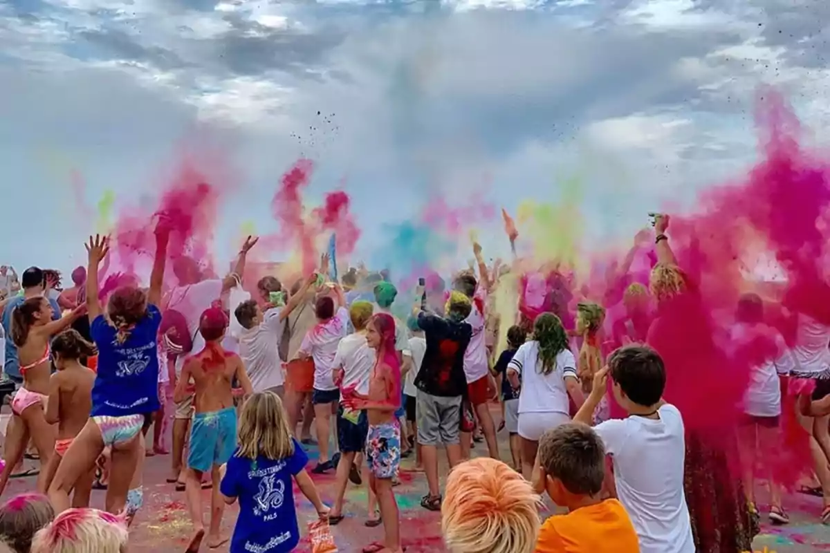 Un grupo de personas, en su mayoría niños, celebran lanzando polvos de colores al aire en un ambiente festivo al aire libre bajo un cielo parcialmente nublado. Un grupo de personas, en su mayoría niños, celebran lanzando polvos de colores al aire en un ambiente festivo al aire libre bajo un cielo parcialmente nublado.