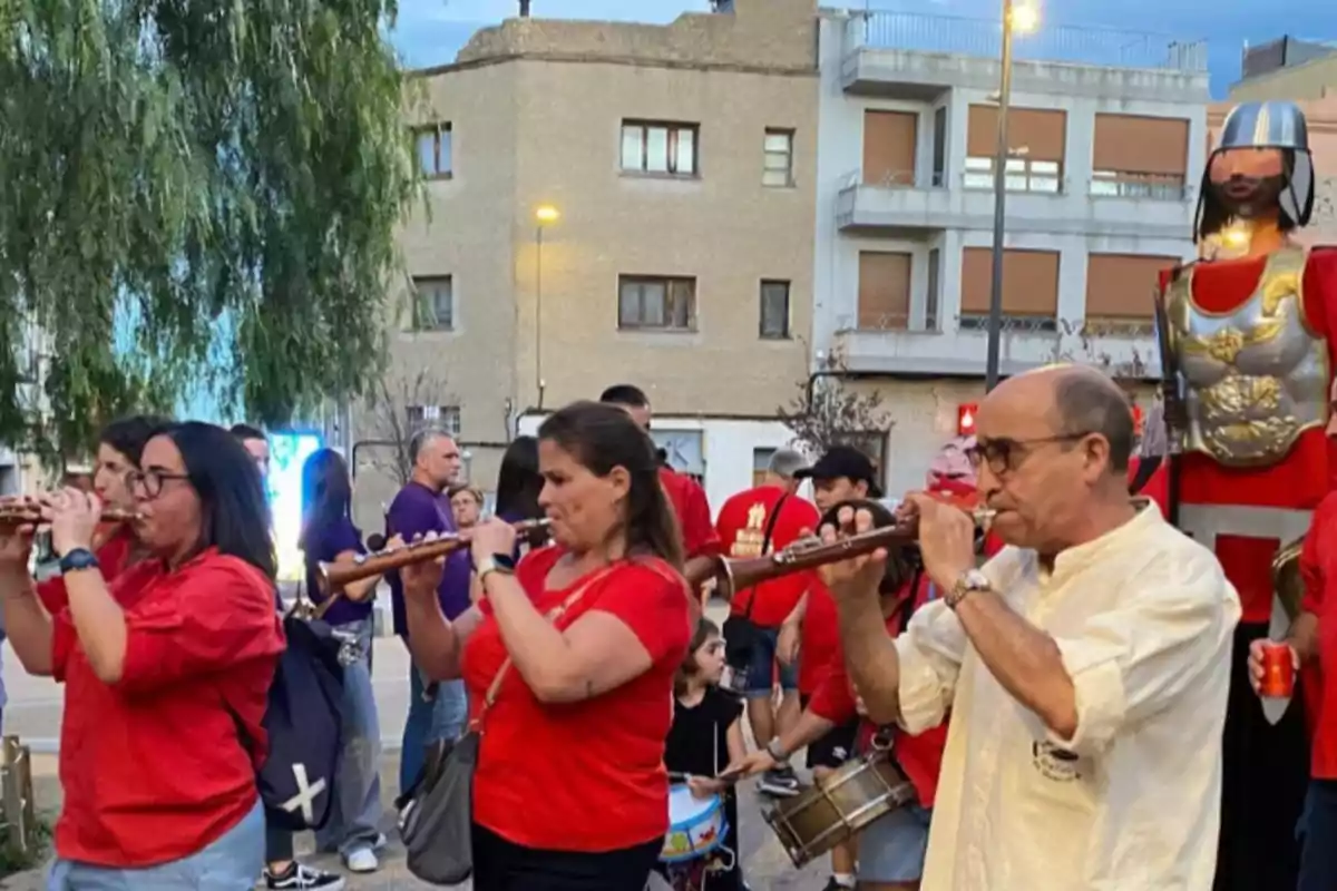 Personas tocando instrumentos de viento en una calle durante una celebración, con un muñeco gigante vestido de caballero al fondo y varias personas observando Personas tocando instrumentos de viento en una calle durante una celebración, con un muñeco gigante vestido de caballero al fondo y varias personas observando