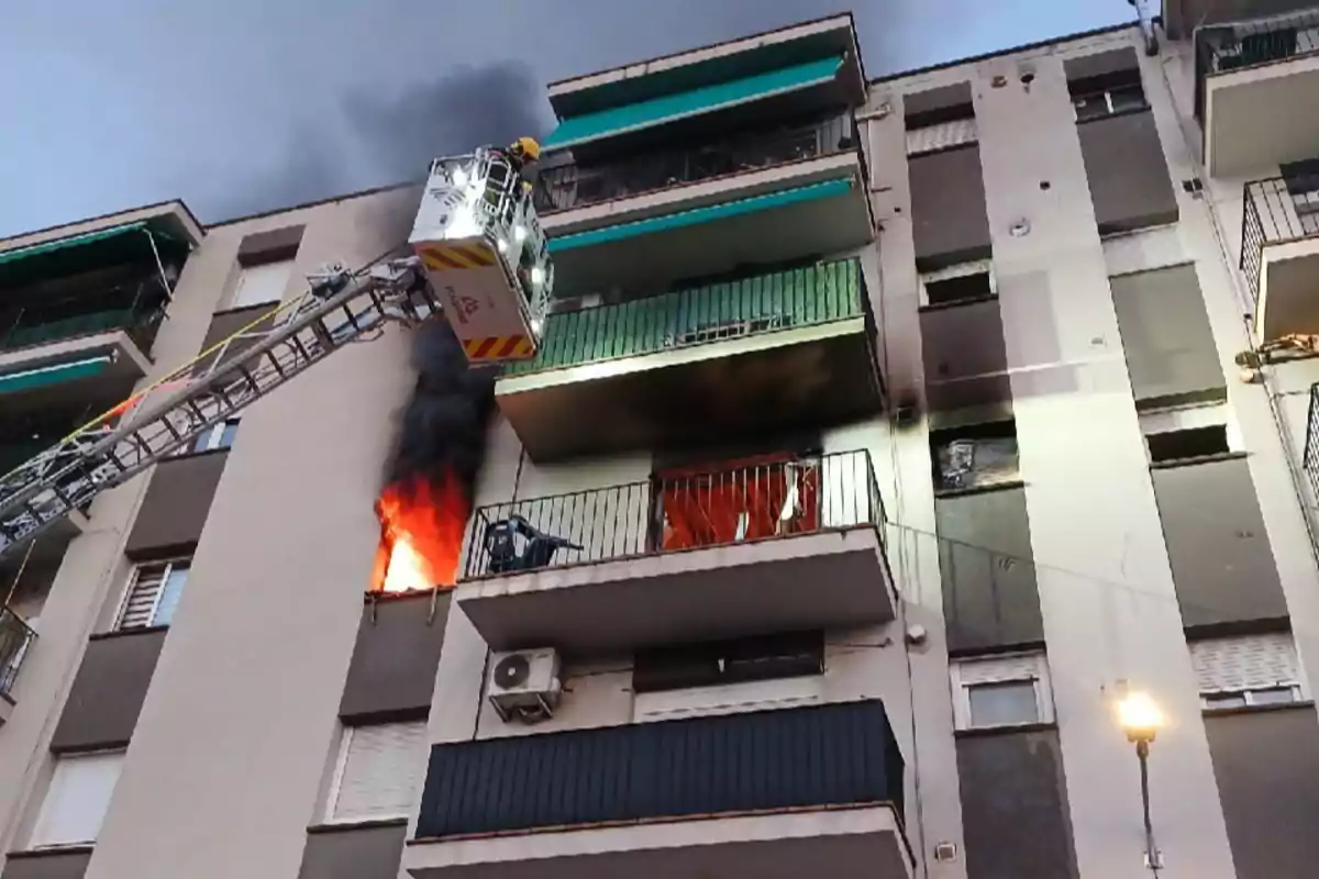Un edifici d’apartaments amb un incendi en un dels pisos mentre un bomber en una grua intenta controlar el foc Un edifici d’apartaments amb un incendi en un dels pisos mentre un bomber en una grua intenta controlar el foc