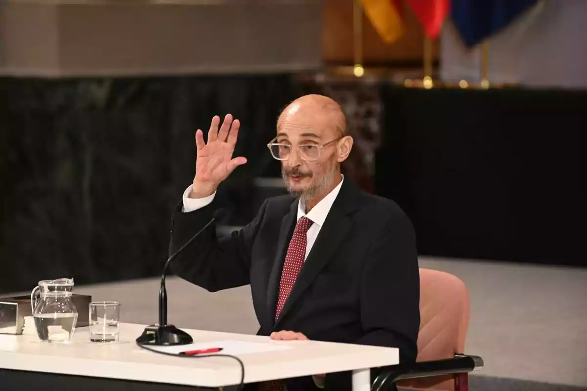 Hombre mayor con gafas y traje oscuro sentado en una mesa blanca levantando la mano junto a un micrófono y una jarra de agua