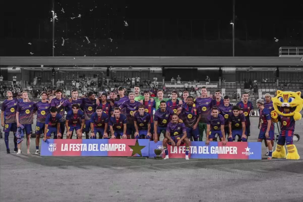 Jugadors d’un equip de futbol posen junts al camp amb el seu uniforme blau i vermell al costat d’una mascota groga i un cartell que diu La Festa del Gamper Jugadors d’un equip de futbol posen junts al camp amb el seu uniforme blau i vermell al costat d’una mascota groga i un cartell que diu La Festa del Gamper