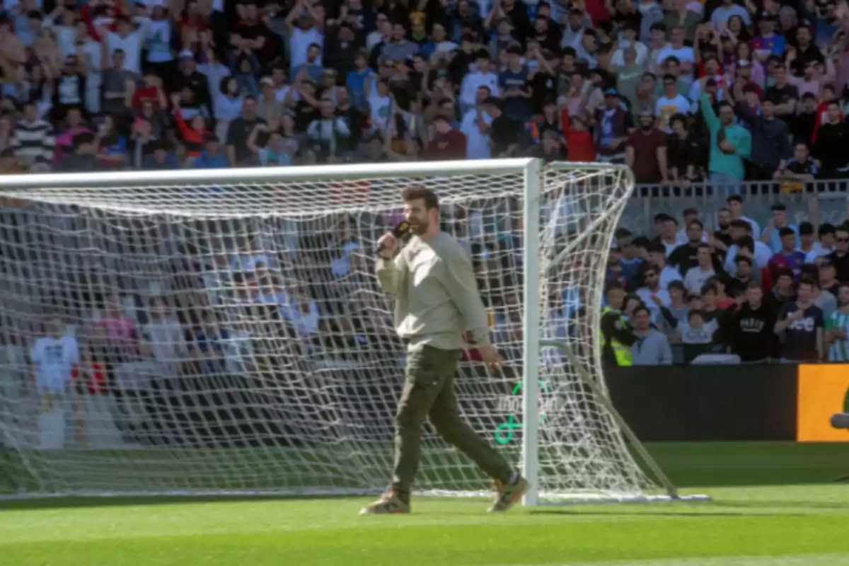 Un home camina davant d'una porteria de futbol mentre sosté un micròfon i una multitud l'observa des de les grades. Un home camina davant d'una porteria de futbol mentre sosté un micròfon i una multitud l'observa des de les grades.