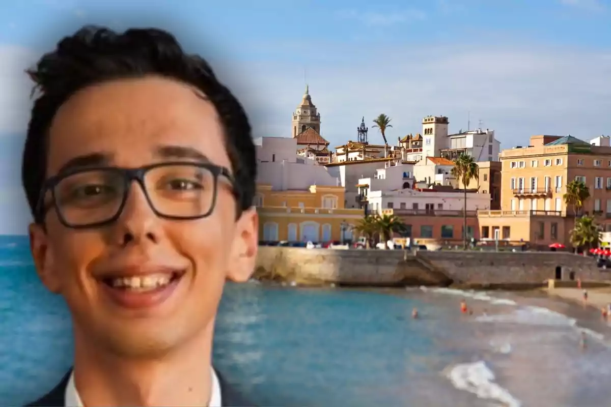 Un home jove amb ulleres somriu en primer pla mentre al fons s’observa una ciutat costanera amb edificis de colors i una platja.