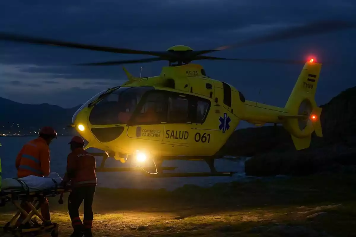 Helicóptero amarillo de emergencias médicas aterrizando de noche mientras dos rescatistas trasladan a una persona en camilla. Helicóptero amarillo de emergencias médicas aterrizando de noche mientras dos rescatistas trasladan a una persona en camilla.