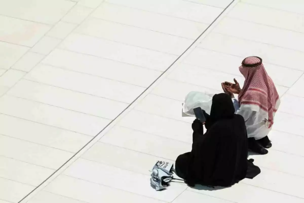 Dos personas con vestimenta tradicional islámica están sentadas sobre un suelo blanco, una de ellas sostiene un teléfono móvil y la otra parece estar rezando. Dos personas con vestimenta tradicional islámica están sentadas sobre un suelo blanco, una de ellas sostiene un teléfono móvil y la otra parece estar rezando.
