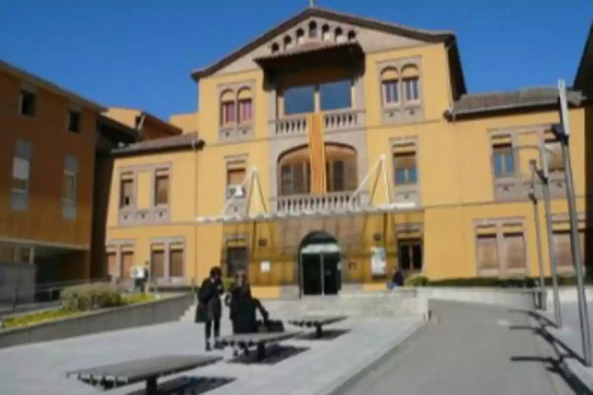 Fachada de un edificio amarillo con detalles arquitectónicos clásicos y una bandera colgada en el balcón central, con dos personas caminando en la plaza frente a la entrada principal bajo un cielo despejado. Fachada de un edificio amarillo con detalles arquitectónicos clásicos y una bandera colgada en el balcón central, con dos personas caminando en la plaza frente a la entrada principal bajo un cielo despejado.