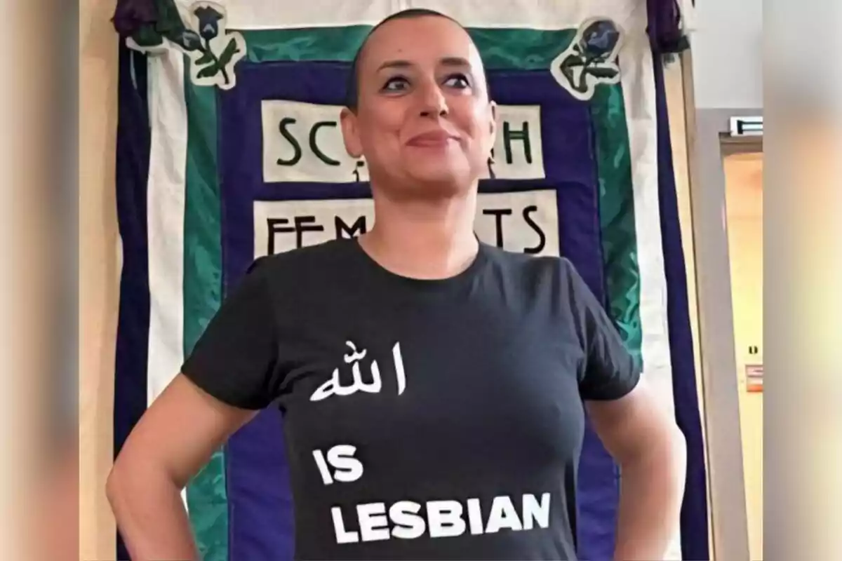 Persona de cabell molt curt portant una samarreta negra amb un missatge provocador en anglès i àrab, dreta davant d’una pancarta feminista.