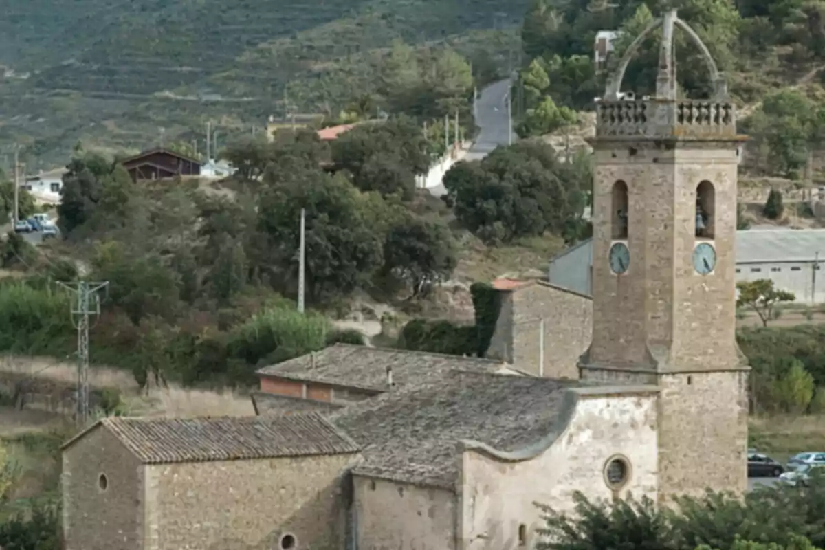 Església de pedra amb campanar i rellotges en un entorn rural envoltat de turons i vegetació Església de pedra amb campanar i rellotges en un entorn rural envoltat de turons i vegetació