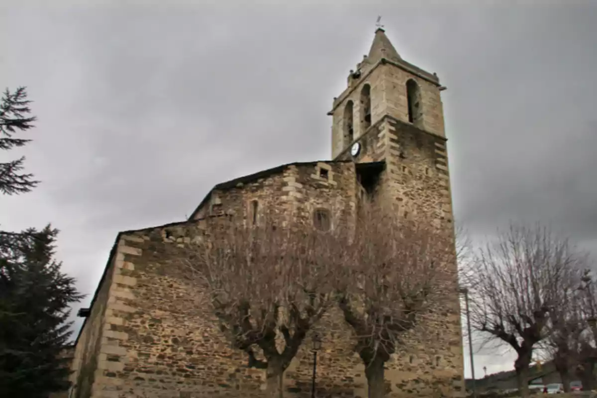 Iglesia de piedra antigua con torre y reloj bajo un cielo nublado rodeada de árboles sin hojas Iglesia de piedra antigua con torre y reloj bajo un cielo nublado rodeada de árboles sin hojas