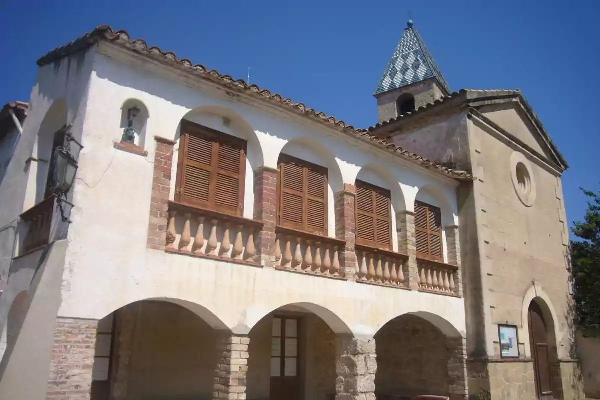 Edifici antic de dues plantes amb arcs a la planta baixa, finestres de fusta al pis superior i una torre amb teulada de mosaic blau sota un cel serè Edifici antic de dues plantes amb arcs a la planta baixa, finestres de fusta al pis superior i una torre amb teulada de mosaic blau sota un cel serè