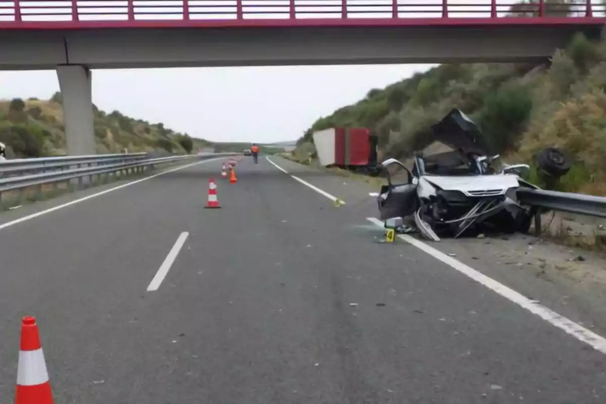 Accidente de tráfico en una autopista con un coche gravemente dañado en la cuneta y un camión volcado, la vía está acordonada con conos y un puente cruza sobre la carretera. Accidente de tráfico en una autopista con un coche gravemente dañado en la cuneta y un camión volcado, la vía está acordonada con conos y un puente cruza sobre la carretera.
