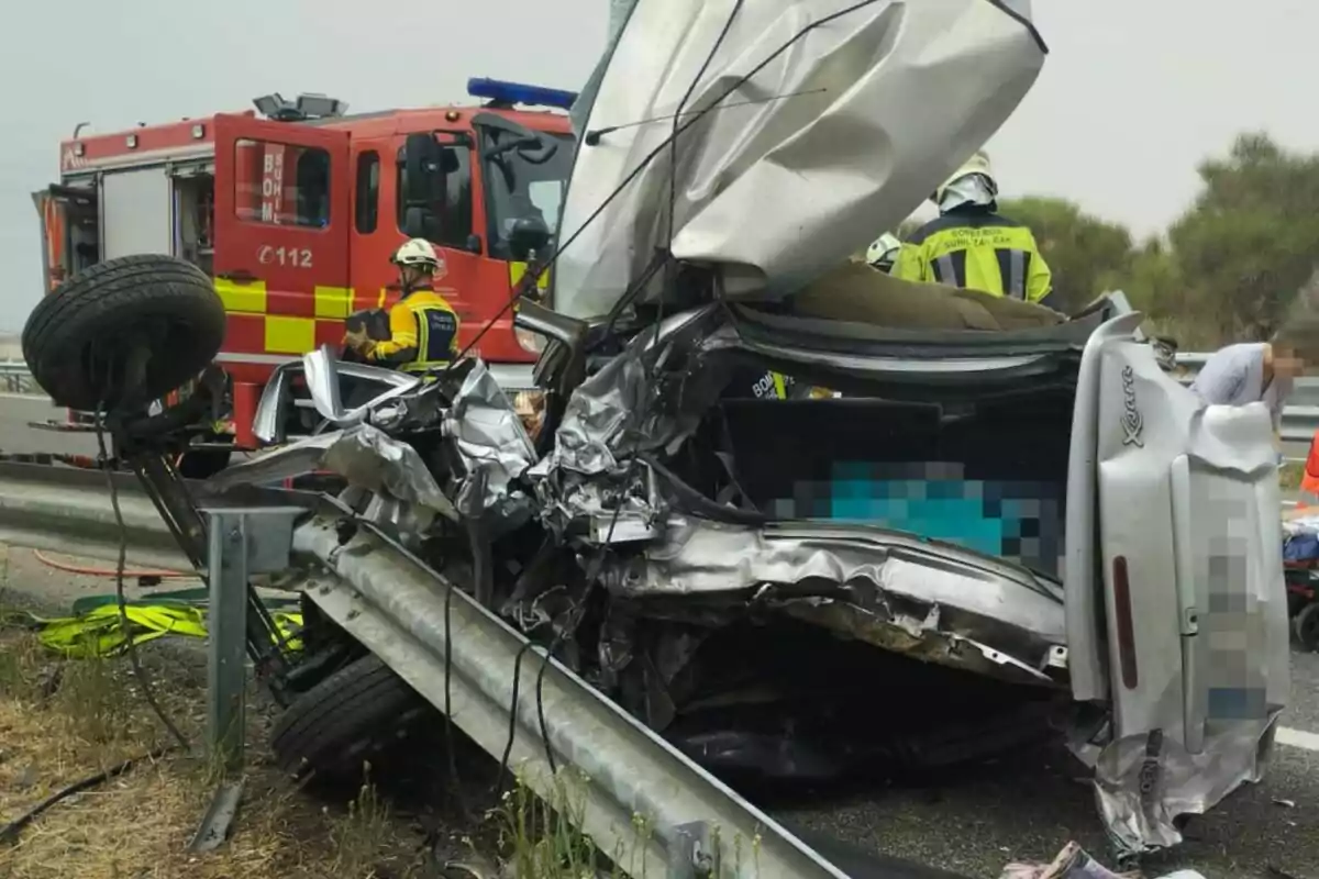 Coche plateado gravemente accidentado contra una valla de seguridad en una carretera con bomberos y un camión de bomberos atendiendo la emergencia