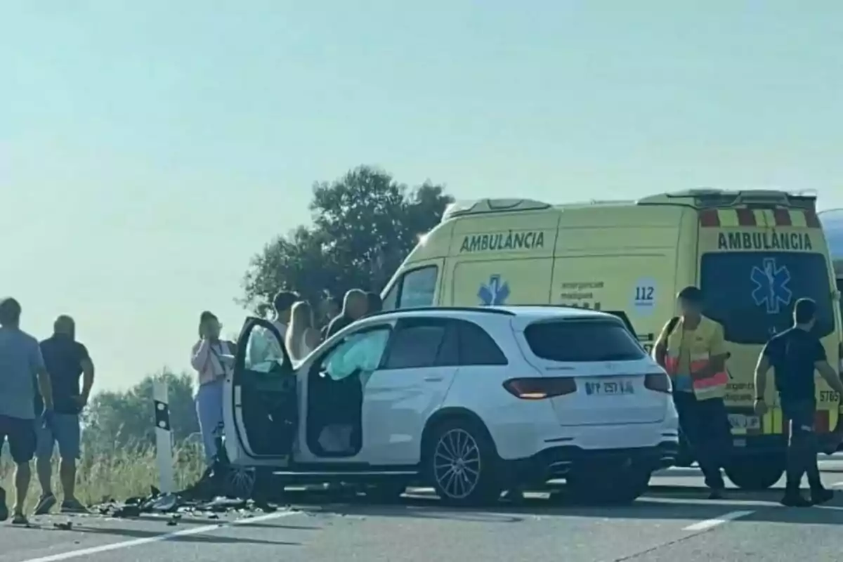 Un cotxe blanc accidentat en una carretera amb la porta oberta, diverses persones al voltant i una ambulància groga estacionada darrere.