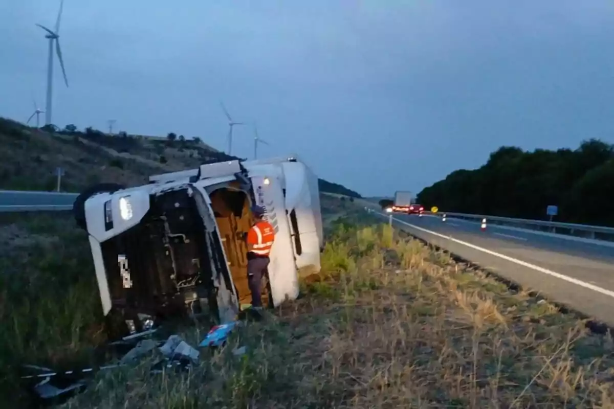 Camió bolcat a la cuneta d’una carretera al capvespre amb un treballador amb armilla reflectant revisant la cabina i diversos aerogeneradors al fons.