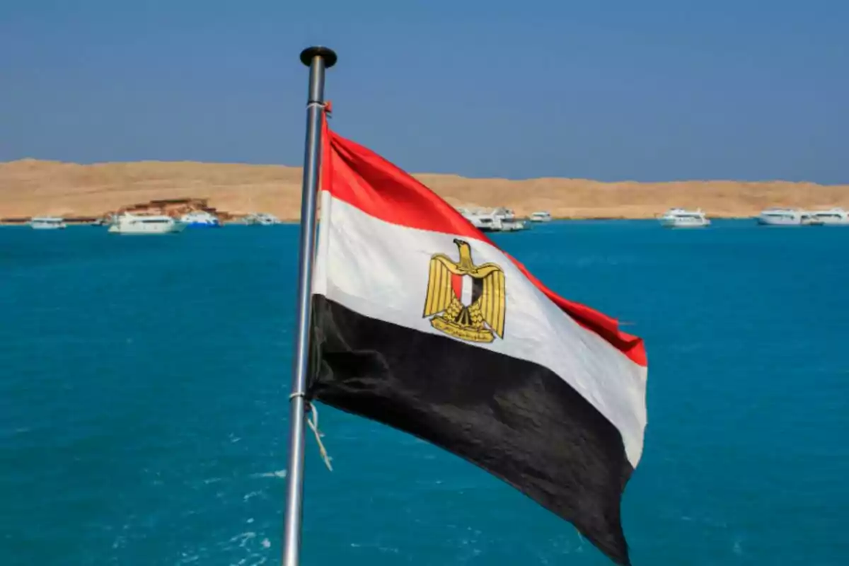 Bandera de Egipto ondeando frente a un mar azul con barcos y un paisaje desértico al fondo Bandera de Egipto ondeando frente a un mar azul con barcos y un paisaje desértico al fondo