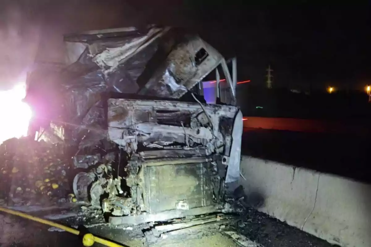 Camió completament calcinat després d’un accident nocturn en una carretera, amb fruites escampades a terra i llums d’emergència al fons