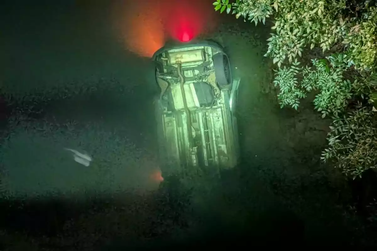 Coche volcado boca abajo sumergido en el agua cerca de la orilla en Galicia con luces traseras encendidas y ramas de árboles al costado. Coche volcado boca abajo sumergido en el agua cerca de la orilla en Galicia con luces traseras encendidas y ramas de árboles al costado.
