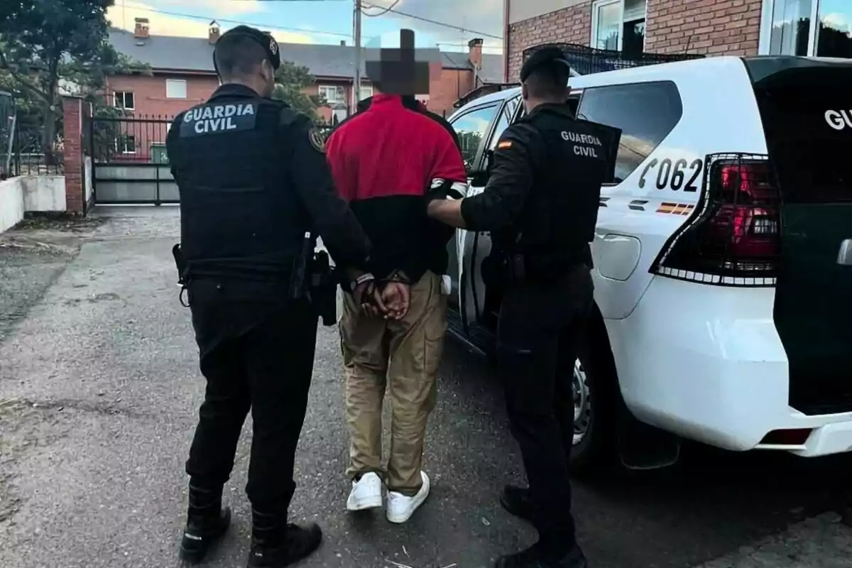 Dos agents de la Guàrdia Civil escorten una persona emmanillada cap a un vehicle oficial en un carrer residencial. Dos agents de la Guàrdia Civil escorten una persona emmanillada cap a un vehicle oficial en un carrer residencial.