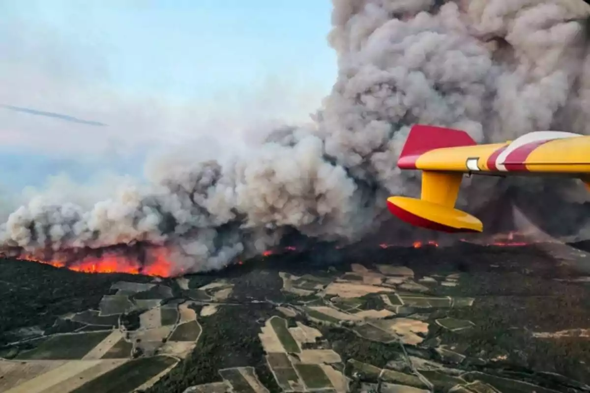 Un avión amarillo y rojo sobrevuela un incendio forestal que produce una gran columna de humo sobre un paisaje de campos y bosques.