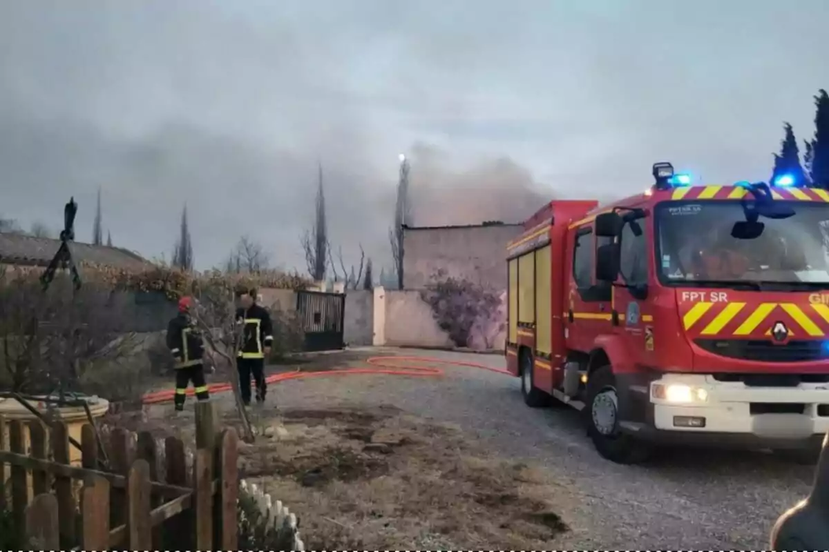 Camión de bomberos con luces encendidas y dos bomberos en una zona residencial donde se observa humo saliendo de una vivienda Camión de bomberos con luces encendidas y dos bomberos en una zona residencial donde se observa humo saliendo de una vivienda