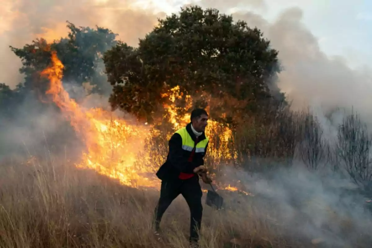 Hombre con chaleco de seguridad intenta controlar un incendio forestal mientras el fuego avanza entre la vegetación y el humo llena el ambiente Hombre con chaleco de seguridad intenta controlar un incendio forestal mientras el fuego avanza entre la vegetación y el humo llena el ambiente