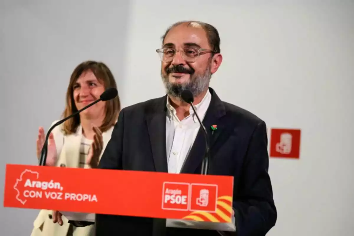Javier Lambán con barba y gafas hablando en un atril del PSOE de Aragón mientras una mujer aplaude al fondo Javier Lambán con barba y gafas hablando en un atril del PSOE de Aragón mientras una mujer aplaude al fondo