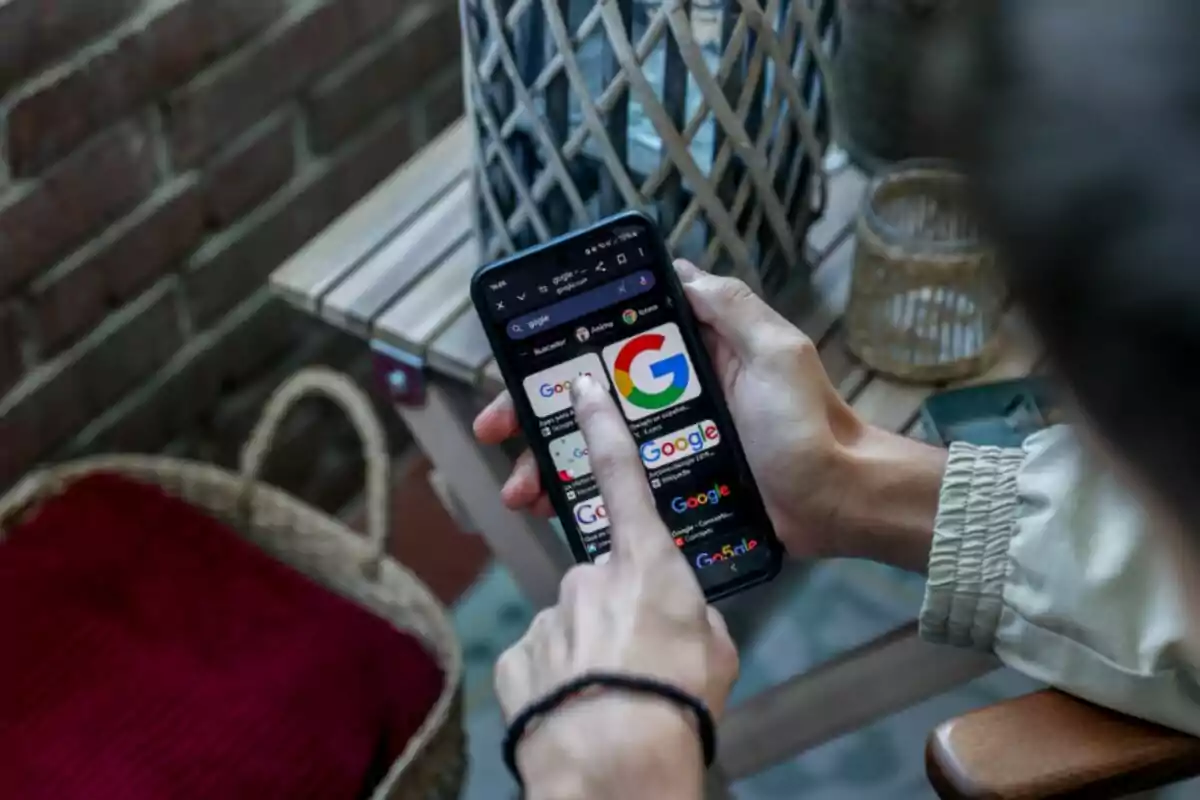 Persona utilitzant un telèfon mòbil per buscar imatges de Google mentre està asseguda en una terrassa amb una taula de fusta i una cistella al costat. Persona utilitzant un telèfon mòbil per buscar imatges de Google mentre està asseguda en una terrassa amb una taula de fusta i una cistella al costat.