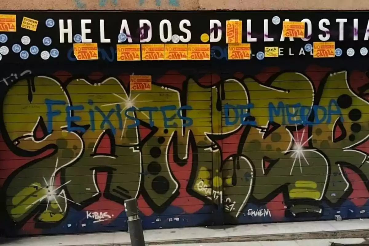 Persiana de una heladería con grafitis de colores, pintadas azules y varios adhesivos amarillos y blancos en la parte superior Persiana de una heladería con grafitis de colores, pintadas azules y varios adhesivos amarillos y blancos en la parte superior