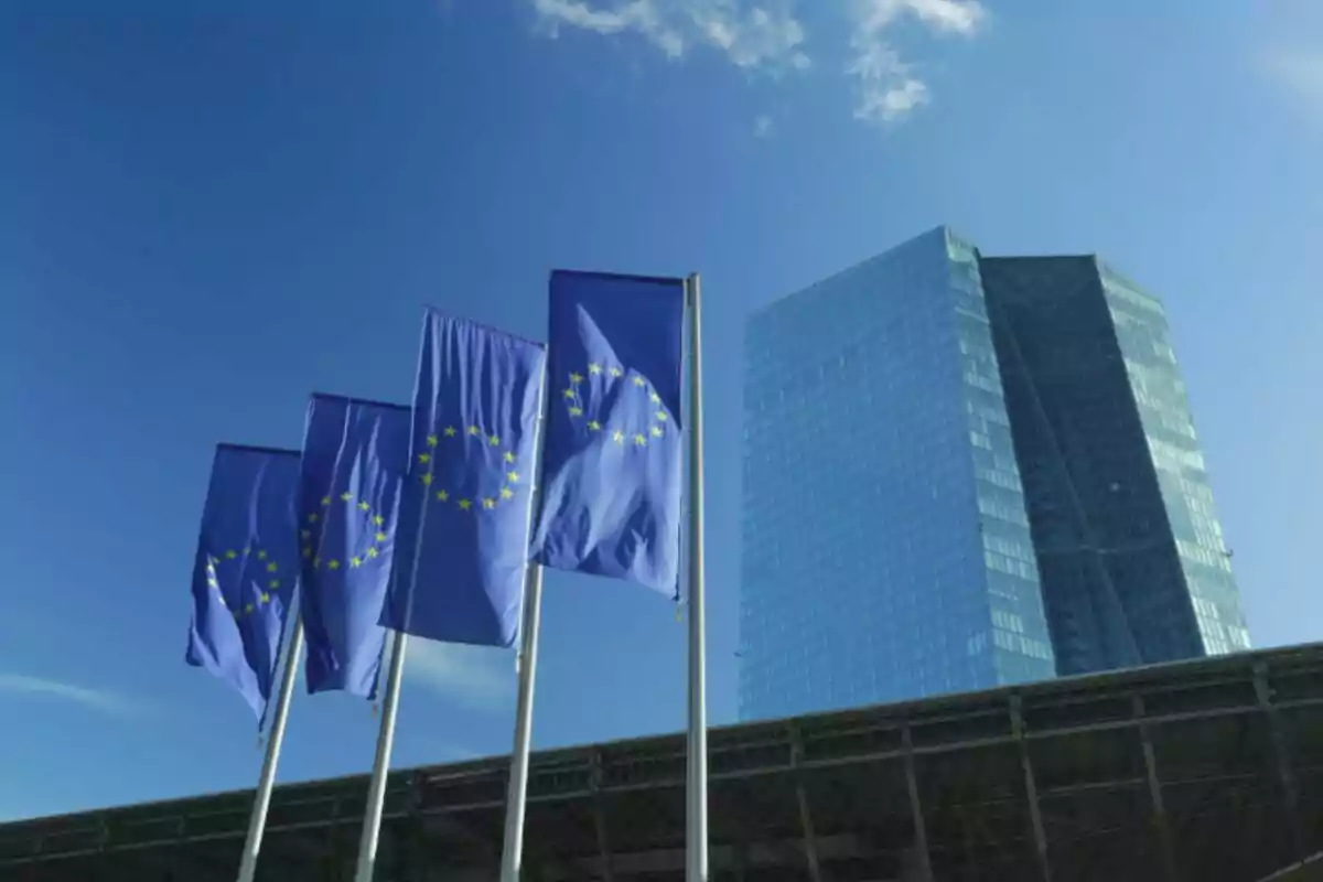 Cuatro banderas de la Unión Europea ondean frente a un edificio moderno de cristal bajo un cielo azul. Cuatro banderas de la Unión Europea ondean frente a un edificio moderno de cristal bajo un cielo azul.