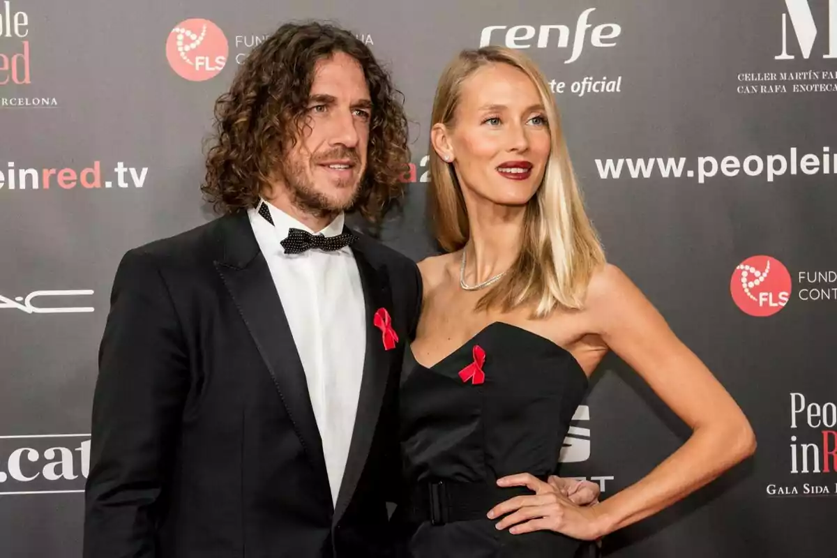 Parella elegantment vestida posant en una catifa vermella amb llaços vermells a la solapa davant d’un fons amb logotips de patrocinadors.