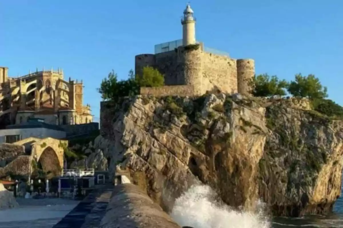 Faro sobre una fortaleza de piedra en lo alto de un acantilado con olas rompiendo en las rocas y una iglesia antigua al fondo bajo un cielo despejado en Cantabria. Faro sobre una fortaleza de piedra en lo alto de un acantilado con olas rompiendo en las rocas y una iglesia antigua al fondo bajo un cielo despejado en Cantabria.
