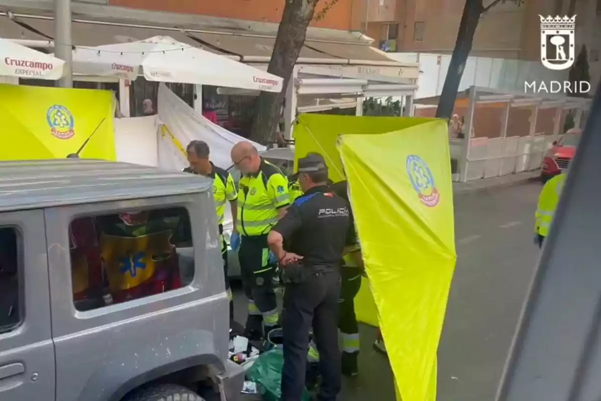 Diversos paramèdics i un policia atenen una emergència al costat d’un vehicle gris en un carrer de Madrid, amb pantalles grogues de protecció i una terrassa de restaurant al fons. Diversos paramèdics i un policia atenen una emergència al costat d’un vehicle gris en un carrer de Madrid, amb pantalles grogues de protecció i una terrassa de restaurant al fons.