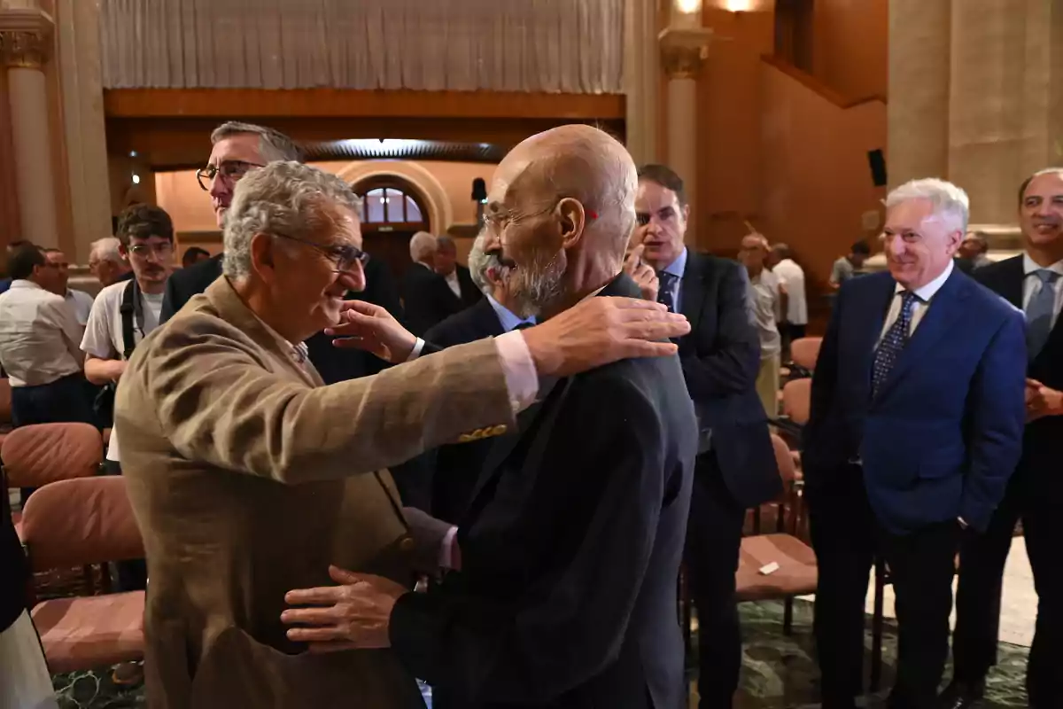 Dos hombres mayores se saludan afectuosamente en un evento formal mientras otras personas observan y conversan en el fondo dentro de un salón elegante. Dos hombres mayores se saludan afectuosamente en un evento formal mientras otras personas observan y conversan en el fondo dentro de un salón elegante.