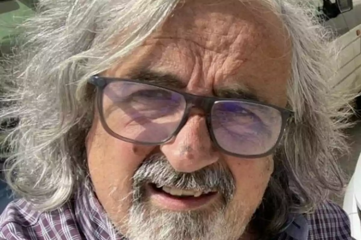 Hombre de cabello canoso y largo con gafas y barba, usando una camisa a cuadros y sonriendo al aire libre