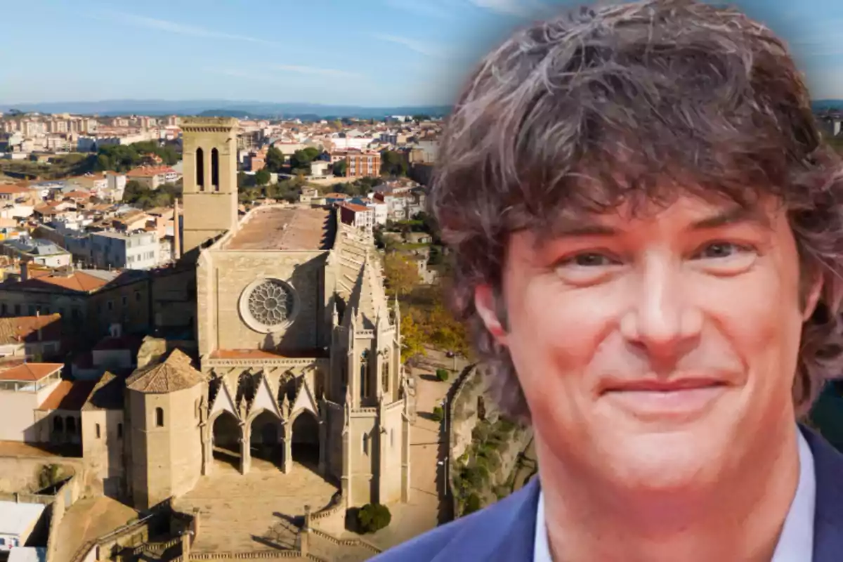 Un home somrient en primer pla amb una església antiga i una ciutat al fons en un dia assolellat