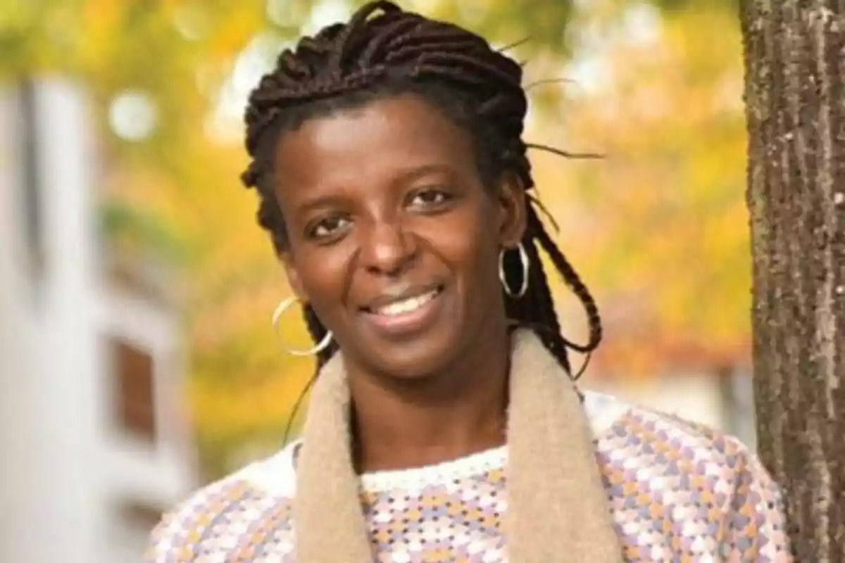 Mujer sonriente con trenzas y aretes de aro posando al aire libre junto a un árbol con fondo de hojas amarillas Mujer sonriente con trenzas y aretes de aro posando al aire libre junto a un árbol con fondo de hojas amarillas