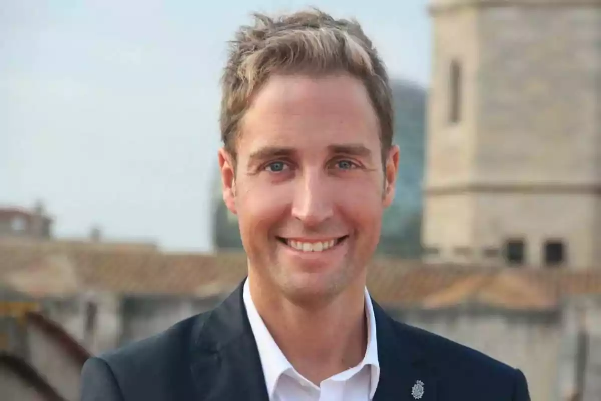 Hombre joven de cabello corto y rubio con saco oscuro y camisa blanca sonriendo al aire libre con edificios de fondo