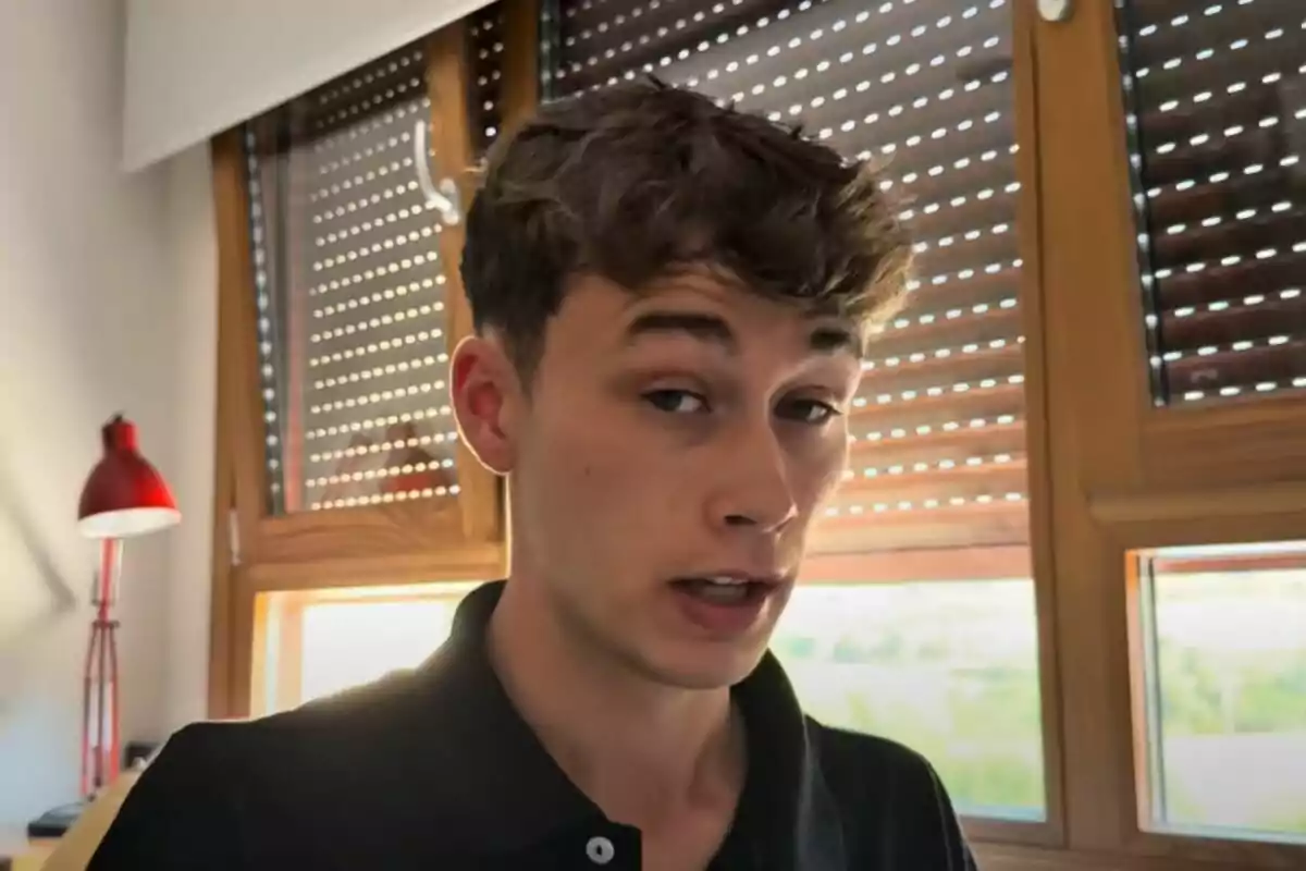 Pla curt de Jorge Rey vestit amb camisa negra parlant de la meteorologia davant d’una finestra amb persianes i una làmpada vermella al fons.